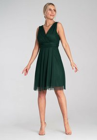 Grünes ärmelloses Kleid mit V-Ausschnitt, enganliegendem Oberteil und schwingendem Rock. Der Stoff ist glatt, mit einer transparenten unteren Schicht für zusätzliche Details.