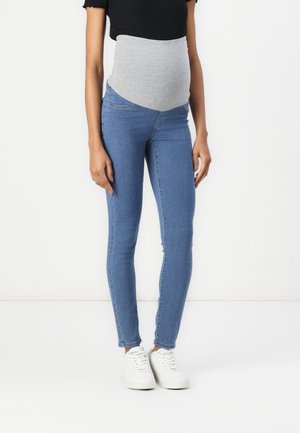 Personne enceinte portant un jean de maternité bleu avec un panneau ventral extensible gris, un haut noir et des baskets blanches, debout sur un fond blanc.