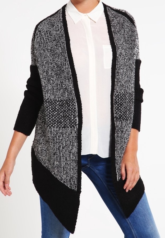 Femme portant un cardigan ouvert texturé noir et gris sur une chemise blanche à col et un jean bleu, debout devant un fond blanc.