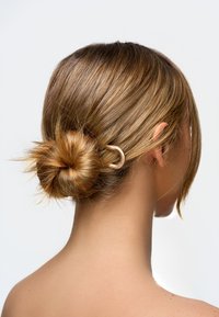 Nainen, jolla on vaaleanruskeat hiukset kiinnitettynä matalaan bun-solmuun kultaisella U-muotoisella hiussoljella, kuvattuna takaapäin yksinkertaista taustaa vasten.