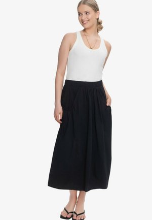 Uśmiechnięta kobieta ubrana w biały top bez rękawów, długą czarną spódnicę z kieszeniami, czarne japonki i złoty naszyjnik, stoi z rękami w kieszeniach.