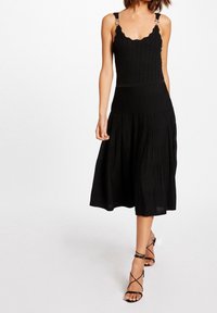 Schwarzes gestricktes Kleid mit gewelltem Ausschnitt, geripptem Oberteil, hoher Taille, schwingendem Faltenrock und verstellbaren Trägern mit metallischen Akzenten.