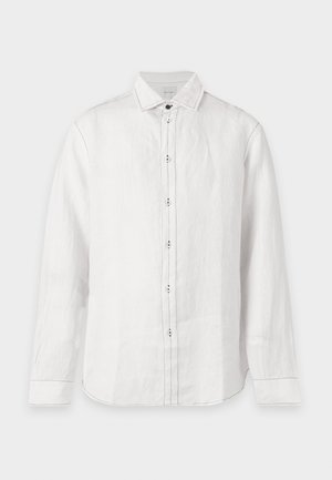Homme debout portant une chemise blanche à manches longues boutonnée, un pantalon noir et des mocassins jaunes sur un fond clair uni.