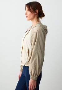 Beige leichte Kapuzenjacke mit gerafften Bündchen, Reißverschluss vorne und lockerer Passform, kombiniert mit navyfarbigen Leggings. Glattes, strukturiertes Material.