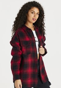 Camicia a quadri rossa e nera in flanella con chiusura a bottoni, due tasche sul petto e una vestibilità leggermente over-size, indossata sopra una maglietta bianca.