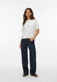 Hvid bluse med korte ærmer og guld accenter, parret med mørkeblå brede jeans. Model står mod en ensfarvet hvid baggrund.