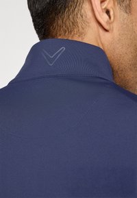 Close-up of man's neck and ear wearing a navy blue collared shirt with a subtle reflective checkmark logo on the back of the collar.