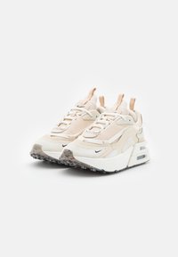 Zapatillas de deporte color beige claro con una parte superior texturizada, que presenta detalles de malla y cuero, suela blanca con amortiguación de aire y acentos negros.