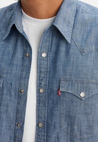 Vaaleansininen farkkupaita, jossa on terävä kaulus, painonapit ja kaksi rintataskua. Kangas näyttää pehmeältä ja teksturoidulta.