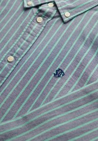 Chemise à rayures bleues et vertes avec boutons blancs et petit logo de poisson brodé en bleu foncé sur la poitrine.