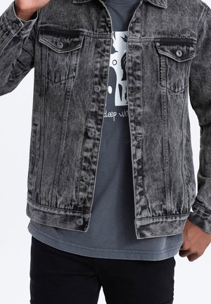 Mężczyzna w rozpiętej wyblakłej czarnej kurtce dżinsowej na ciemnym t-shircie z grafiką i czarnych spodniach, pozuje z uniesioną jedną ręką.