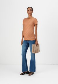 Lys brun kortermet topp med et diamantmønster, kombinert med blå utsvungne jeans og svarte sandaler, holder en beige bag.