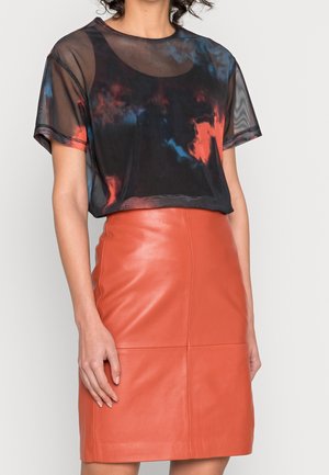 Frau trägt ein durchsichtiges schwarzes und mehrfarbiges kurzärmeliges Oberteil, kombiniert mit einem hoch taillierten orangen Lederrock vor einem einfarbigen Hintergrund.