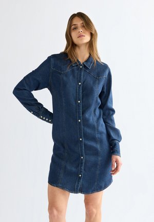 Vrouw met lang haar die een donkerblauwe denim overhemdjurk met knopen en lange mouwen draagt, staand tegen een effen lichte achtergrond.