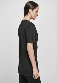 Schwarzes Baumwoll-T-Shirt, kurze Ärmel, leicht elongated Form, mit einem pinken Grafikdesign auf der Vorderseite, glatte Textur, keine sichtbaren Hardware.