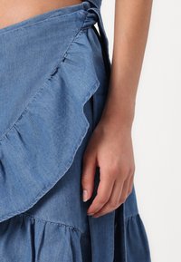Denim zavita krila z nagubanjem na robu, zavezovanjem in večplastno zasnovo. Tkanina je svetlo do srednje modre barve z gladko teksturo.