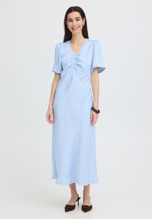 Femeie zâmbitoare, îmbrăcată într-o rochie midi bleu deschis cu mâneci bufante, cu decolteu în V cu pliuri, purtând o brățară neagră și pantofi negri cu vârf ascuțit, stând pe un fundal alb.