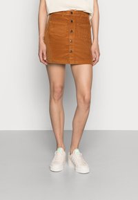 Jupe mini en velours côtelé marron avec des boutons sur le devant et deux poches, associée à des baskets blanches avec des accents pastel. Texture lisse, design ajusté.