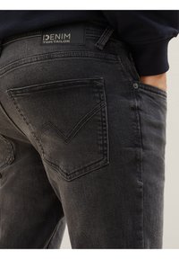 Tmavě šedé džínové kalhoty s černou nálabelkou "DENIM TOM TAILOR." Vyznačují se jedinečným prošitím zadní kapsy. Měkká, strukturovaná textilie.