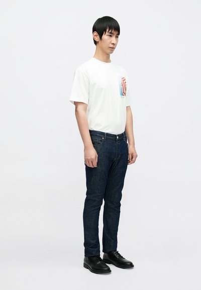 T-shirt blanc à manches courtes avec une poche graphique colorée, associé à un jean en denim bleu foncé et des chaussures noires. Design simple et coupe ajustée.