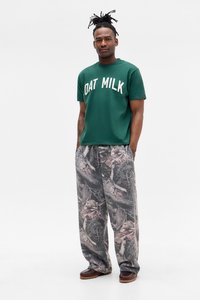 Green cotton T-shirt with "OAT MILK" text. Loose-fit gray pants with a camouflage pattern. Brown shoes with a textured sole.