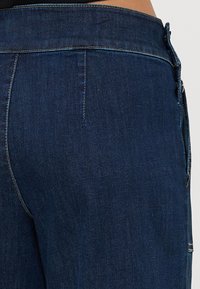 Los jeans de mezclilla azul oscuro presentan una cintura de talle medio, bolsillos traseros y costuras visibles. La tela tiene una textura suave con un ligero estiramiento.