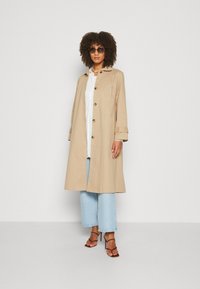 Trench coat bej cu închideri cu buton, mâneci lungi și croi drept, asortat cu blugi tip wide-leg de culoare albastră deschisă și sandale cu toc subțire maro.