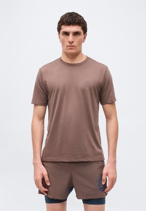 Mężczyzna w brązowej koszuli z krótkim rękawem i pasujących spodenkach stoi na tle jasnego, jednolitego tła.