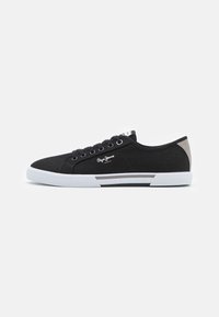 Black canvas sneakers feature white rubber sole, round toe, and logo detailing. Back has a gray accent panel. Laced design with six eyelets.