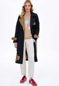 Czarny płaszcz typu trencz z beżowymi akcentami, guzikami i wzorem w panterkę. Do tego beżowy sweter z kolorowymi wzorami grzybów oraz białe spodnie.