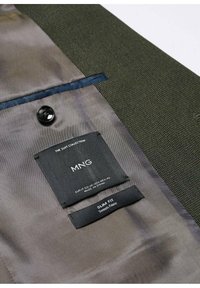 Vue intérieure d'une veste de costume vert foncé montrant un bouton noir, une poche intérieure et deux étiquettes en tissu indiquant la marque MNG et un tissu stretch slim fit.