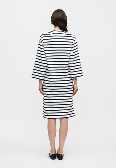 Γυναίκα που φοράει λευκό φόρεμα μήκους μέχρι το γόνατο με μαύρες οριζόντιες ρίγες, στέκεται με την πλάτη στην κάμερα και μαύρα επίπεδα παπούτσια σε λευκό φόντο.