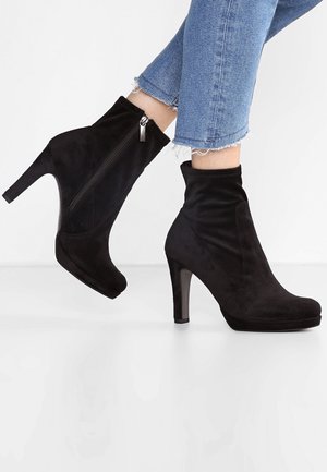 Bottines en daim noir avec un haut talon aiguille, dotées d'une fermeture éclair latérale et d'une texture douce, portées avec un jean bleu à ourlet effiloché.