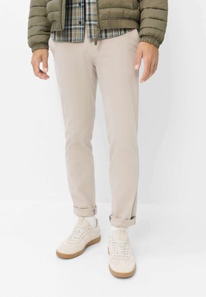 Mann trägt beige Hose, die am Knöchel hochgekrempelt ist, weiße Turnschuhe, ein kariertes Hemd und eine olivgrüne Steppjacke, steht vor weißem Hintergrund.