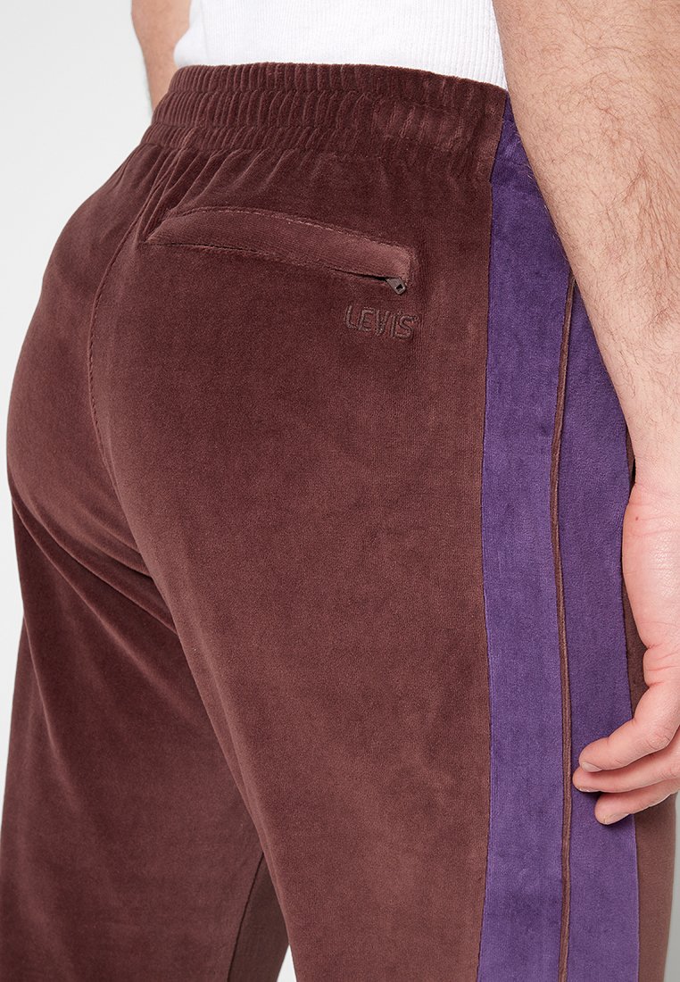 Joggers en velours marron avec une coupe slim et une bande latérale violette ; taille élastique et petite poche zippée à l'arrière. Logo "Levi's" visible.