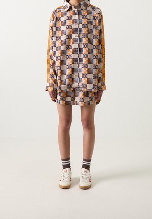 Osoba nosząca dopasowaną pomarańczową, brązową i beżową koszulę z geometrycznym wzorem na guziki oraz szorty, białe trampki i paskowane skarpetki.