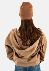 Persoon met lang bruin haar, die een bruine beanie, een bijpassende bruine hoodie en blauwe spijkerbroek draagt, van achteren gefotografeerd tegen een witte achtergrond.