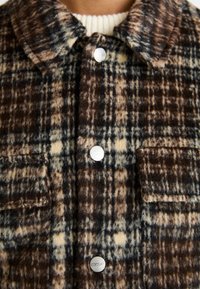 Braun-beige kariertes Hemd-Jacke aus weichem Stoff, ausgestattet mit silbernen Druckknöpfen und zwei Fronttaschen mit feiner Textur.