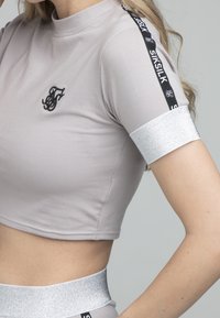 Top corto gris con mangas cortas, que presenta un logo bordado en negro y detalles plateados brillantes en los puños y la cinturilla.