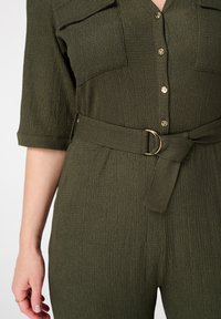 Combinaison olive en tissu texturé, dotée de deux poches poitrine, de fermetures à bouton et d'une ceinture assortie avec une boucle en or.
