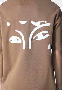 Brun T-shirt med et udskåret design af hvide styliserede øjne på ryggen. Blødt stof med en afslappet pasform og korte ærmer.