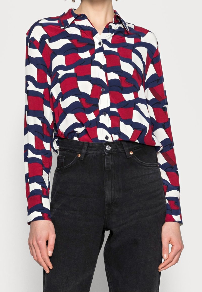 Chemise à manches longues boutonnée avec un motif géométrique ondulé rouge, marine et blanc, rentrée dans un jean noir taille haute.