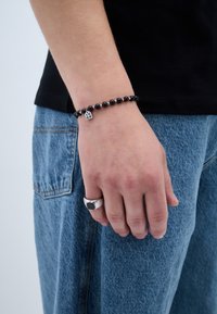 Brățară din mărgele negre cu charm-uri din litere accentuate cu argint, purtată pe o mână. Mâna mai afișează și un inel masiv din argint cu o piatră neagră.