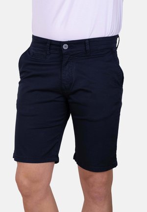 Hombre con pantalones cortos negros hasta la rodilla y una camisa blanca sobre un fondo liso y claro.