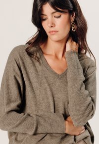 Olijfgroene V-hals trui, gemaakt van zacht gebreid materiaal. Heeft geribbelde manchetten en een relaxte pasvorm. Het model raakt voorzichtig haar nek aan.