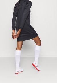 Musta pitkähiha dressi, jossa on teksturoitu kuosi, yhdistettynä valkoisiin polvipitkiin sukkiin ja valkoisiin jalkapallokengiin, joissa on punaisia korostuksia ja logo.