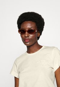 Gafas de sol de tono marrón con lentes rectangulares estrechos, con un acabado brillante, llevadas con una camiseta crema que muestra un logo en relieve.