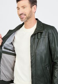 Donkergroene leren jas met een rits aan de voorkant, drukknoopkraag en binnenzakken. Draagt over een lichtgekleurde gebreide trui.