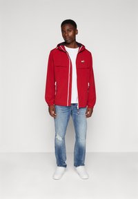 Giacca a vento rossa con zip frontale, due ampie tasche sul petto e cappuccio. Indossata sopra una maglietta bianca e abbinata a jeans blu chiaro. Sneakers bianche.