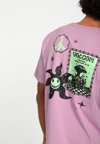 Roze katoenen t-shirt met zwarte grafische prints, waaronder een glimlachend personage, kettingontwerp en tekst. Het ontwerp aan de achterkant bevat een ingelijste illustratie.